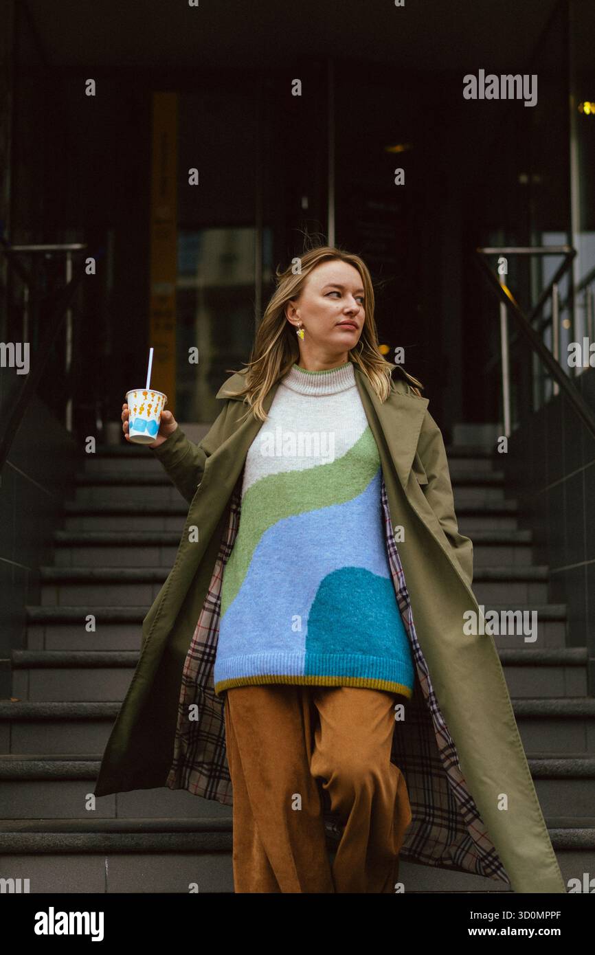 Donna elegante che regge un drink sui gradini urbani indossando un trench all'oliva Foto Stock
