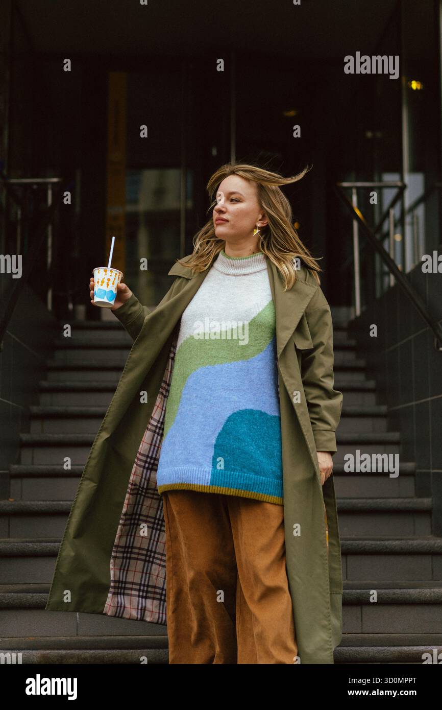 Donna elegante che regge un drink sui gradini urbani indossando un trench all'oliva Foto Stock