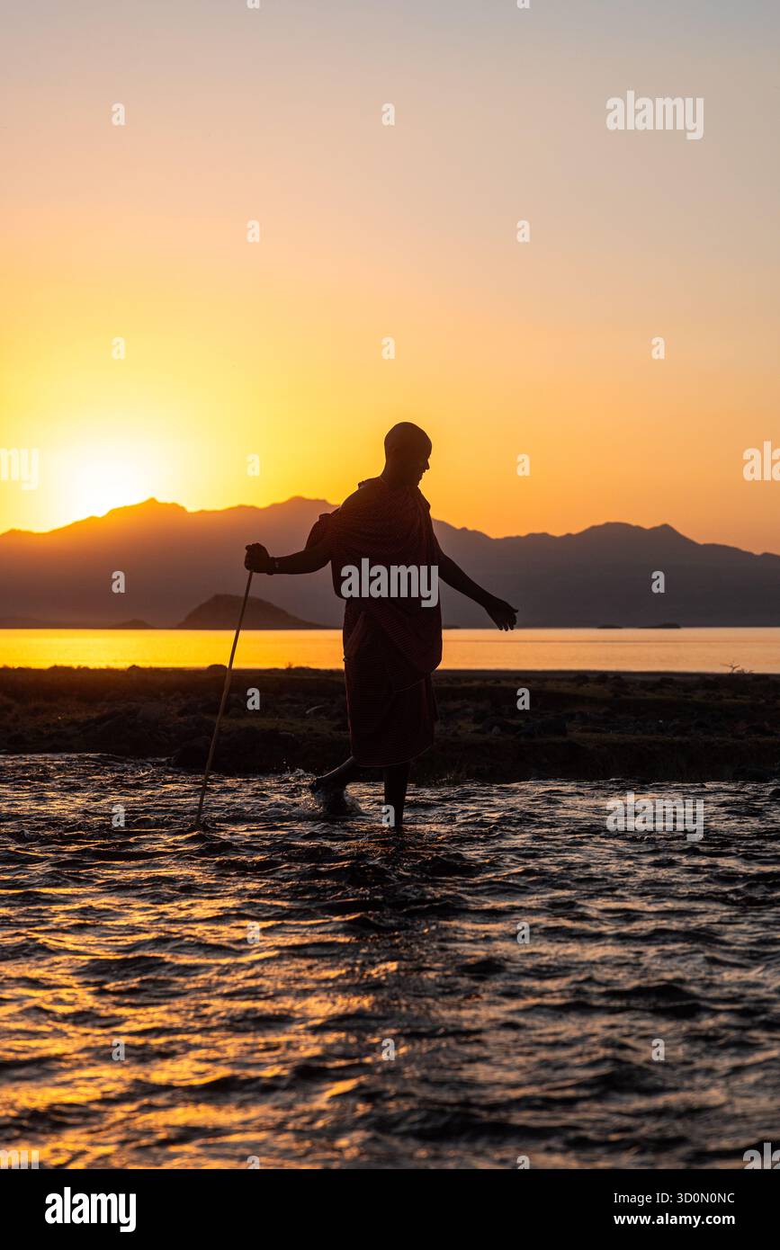 Vista di una figura sagomata che si tuffa attraverso acque poco profonde, tenendo un bastone mentre il sole tramonta dietro montagne lontane, gettando un bagliore dorato, lago Natron, Maasai, Tanzania. Foto Stock