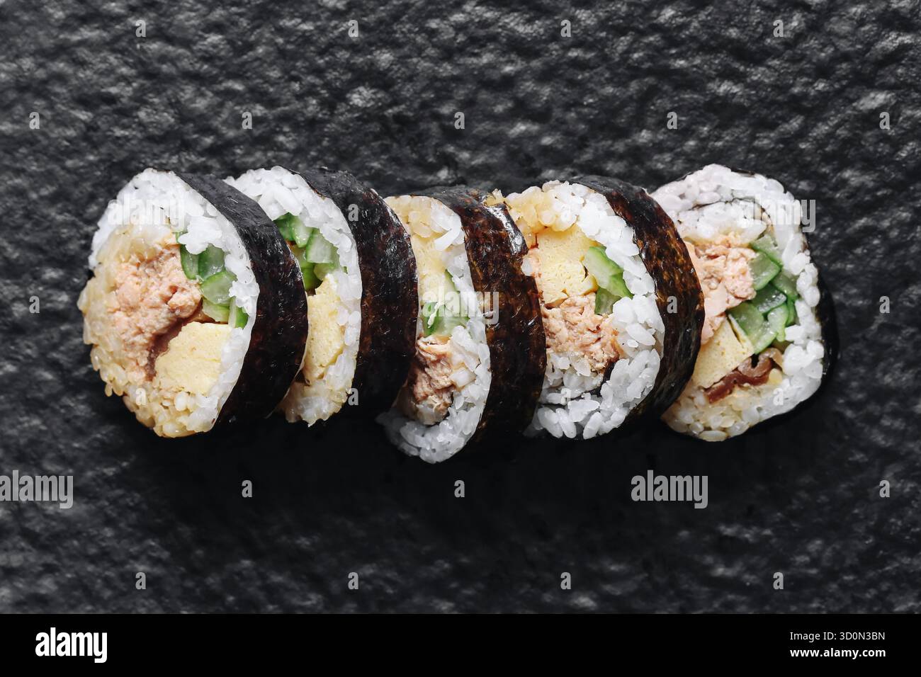 Sushi roll disposti in fila su sfondo in pietra nera. Sushi giapponese maki con nori di riso e ripieno di pesce. Composizione minima degli alimenti. Cugine asiatica. Foto Stock