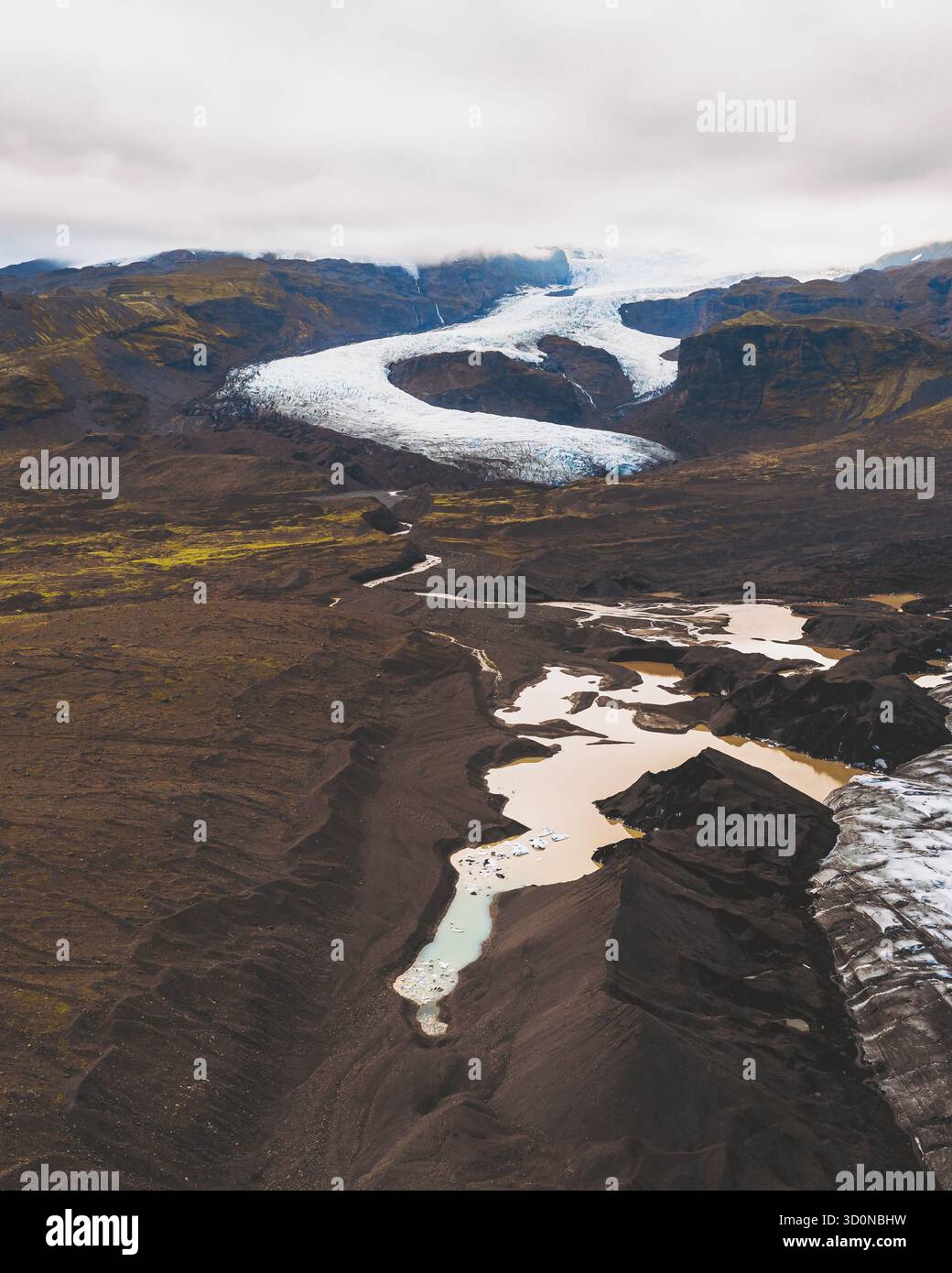 Vista aerea del ghiaccio glaciale che si intreccia attraverso terreni scuri e accidentati, contrastando con il cielo pallido sopra Fjallsárlón, Sveitarfélagið Hornafjörður, Islanda. Foto Stock
