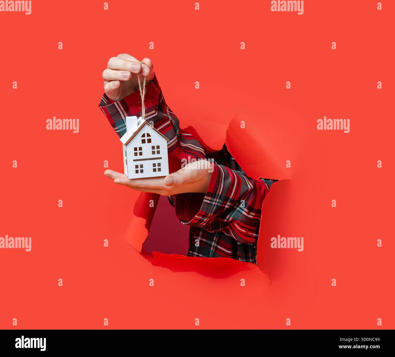 Buon Natale. Le mani reggono un piccolo ornamento bianco in legno per la casa di Natale con una corda attraverso la carta rossa strappata. Foto Stock
