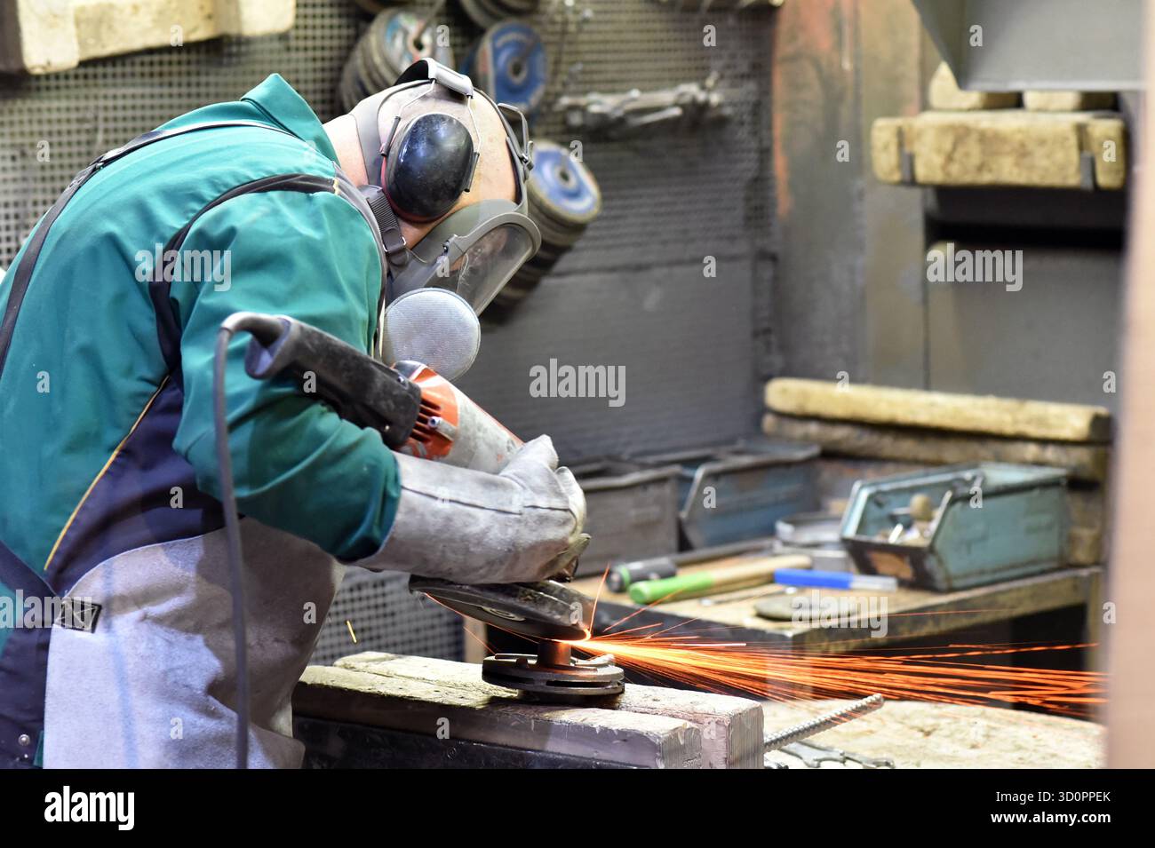 Il lavoratore con una rettificatrice lavora un pezzo in acciaio in un'azienda industriale - sicurezza sul lavoro con protezione per orecchie e occhi - foto ravvicinata Foto Stock