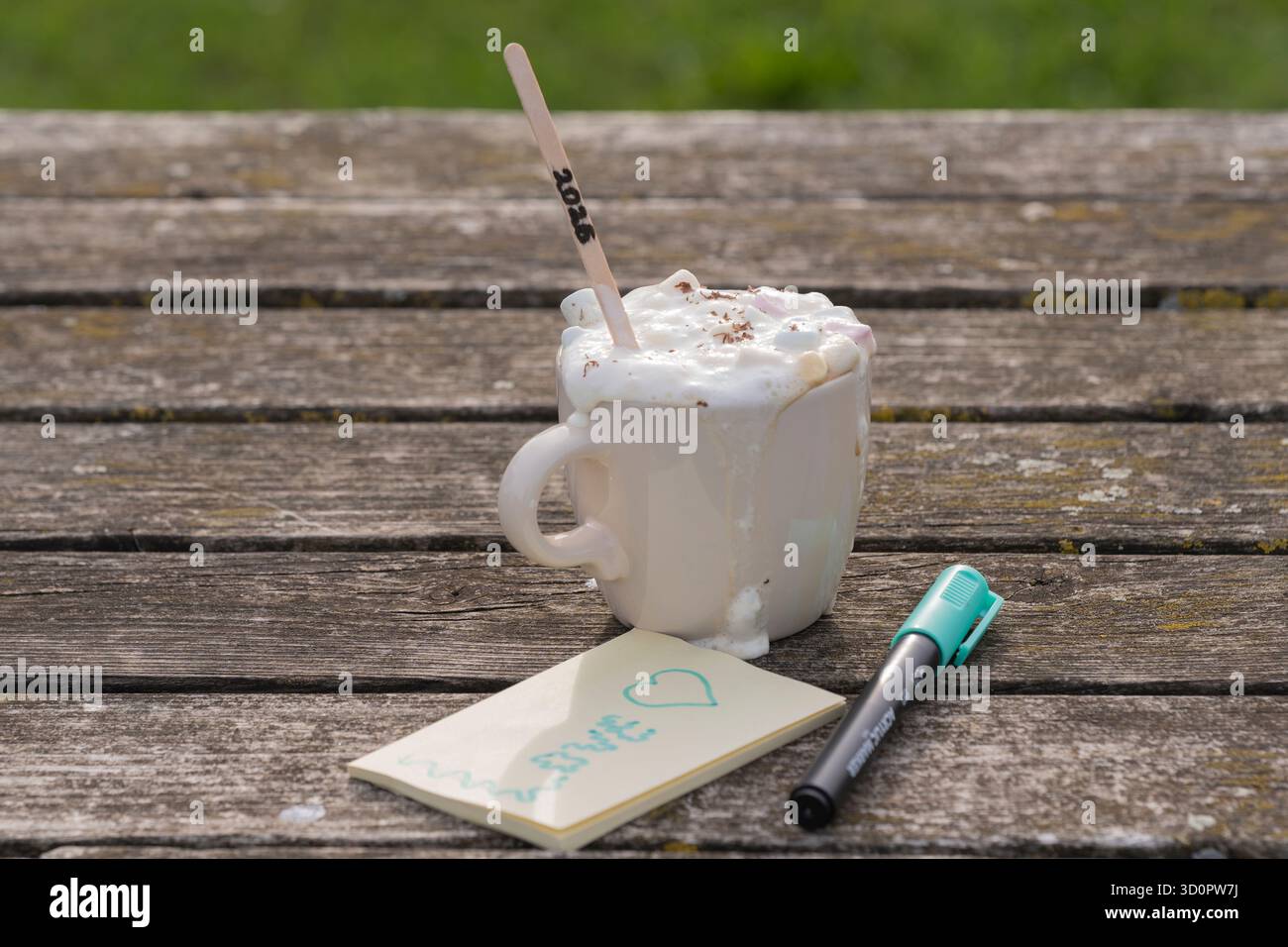 Tazza da caffè bianca con panna montata, marshmallow e bastoncino di legno "2026" accanto al taccuino con cuore e "amore" sul vecchio tavolo di legno - vista laterale Foto Stock
