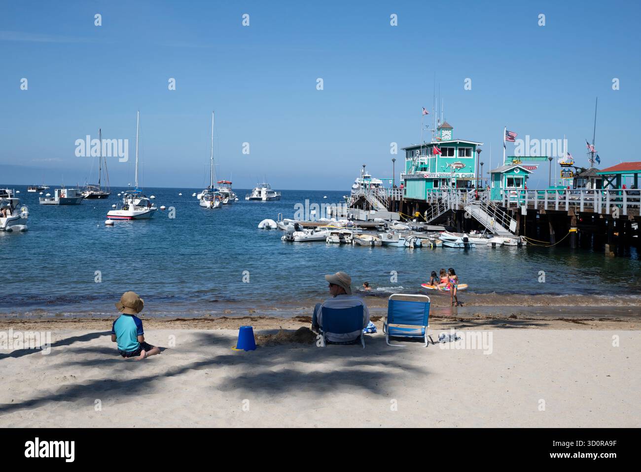Avalon, Catalina Island, CA USA - 25 aprile 2019: Famiglia seduta sulla sabbia a Middle Beach che guarda al Green Pleasure Pier. Foto Stock