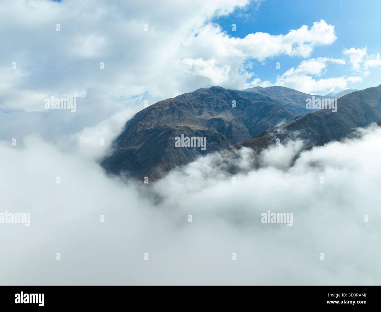 Vista aerea alta di un'aspra e scura vetta di montagna che emerge da uno spesso e vorticoso strato di nuvole bianche o nebbia sotto un cielo azzurro luminoso e frastagliato Foto Stock