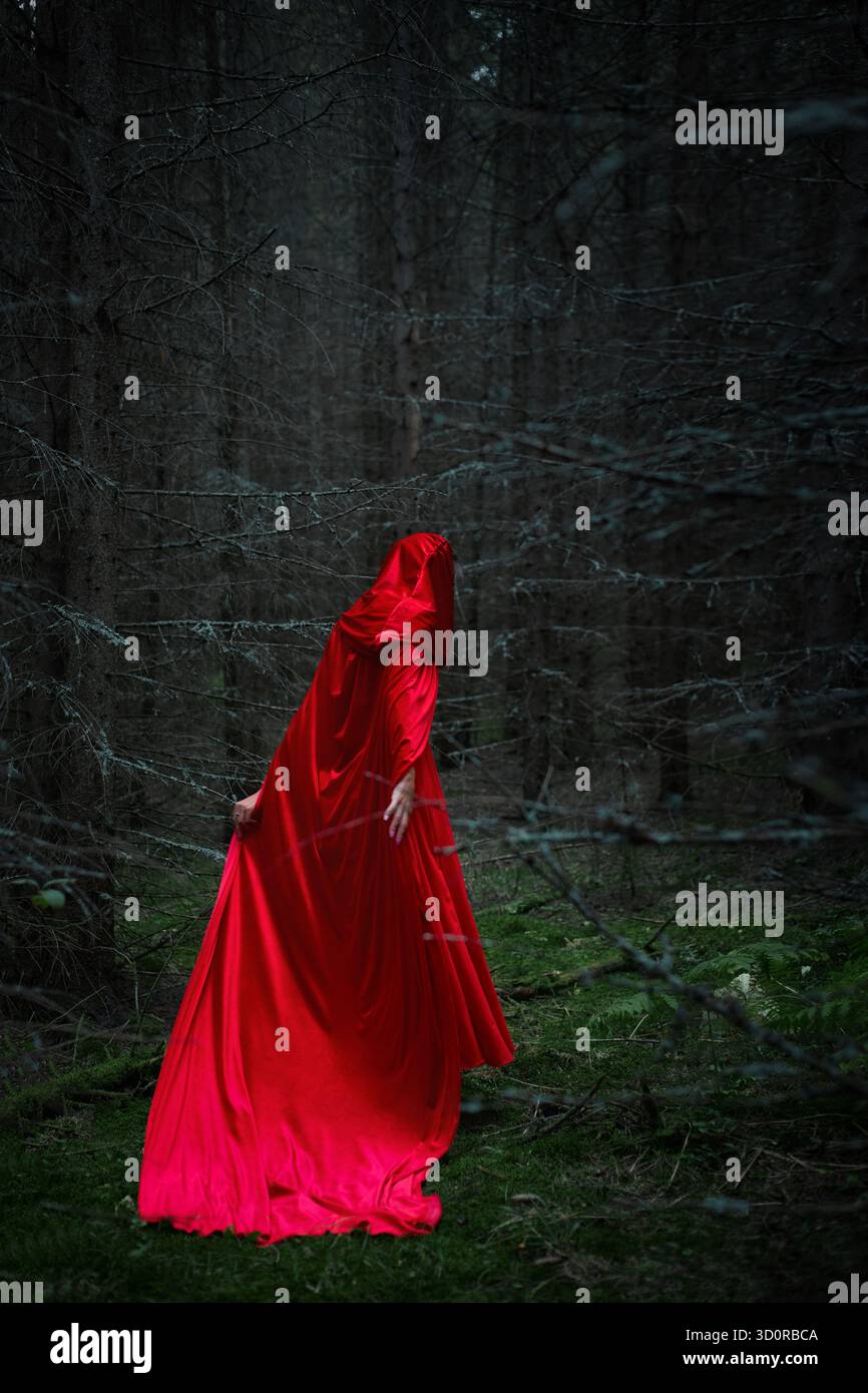 una donna misteriosa vestita con un mantello rosso vibrante vaga attraverso una fitta e buia foresta al crepuscolo. l'atmosfera e' inquietante ma incantevole, circondata da alti tetti Foto Stock