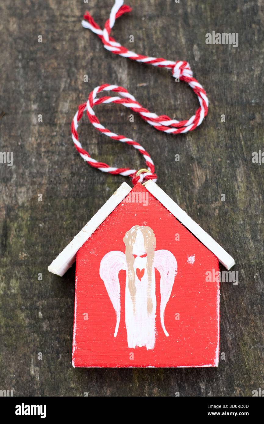 Un affascinante ornamento natalizio fatto a mano caratterizzato da una casa rossa dal design di un angelo bianco, impreziosito da una festosa spago rosso e bianco su legno. Foto Stock