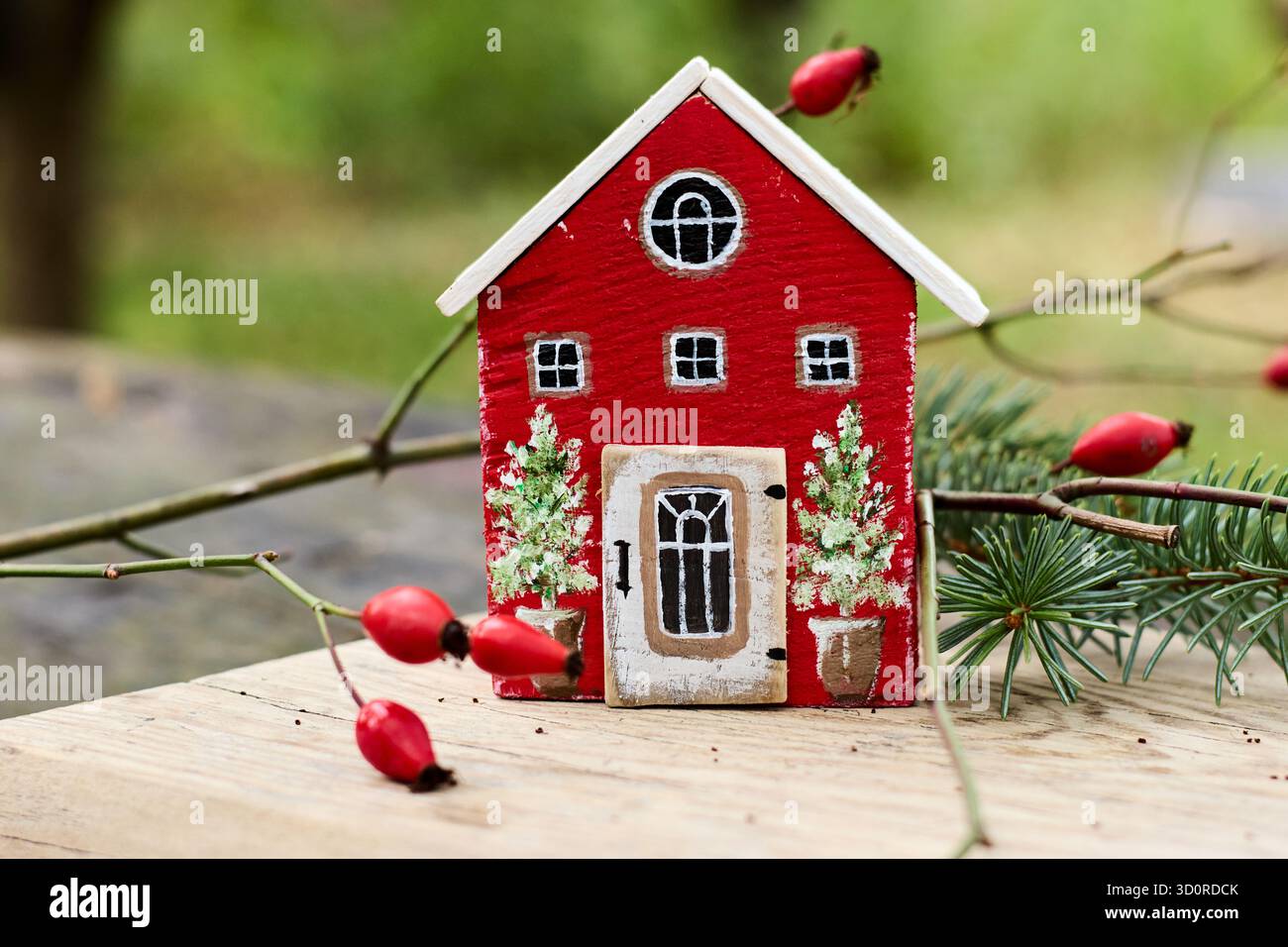 Affascinante casa rossa in miniatura con rifiniture bianche su una superficie in legno, circondata da rami con rosoni fianchi, che catturano una scena stravagante. Foto Stock