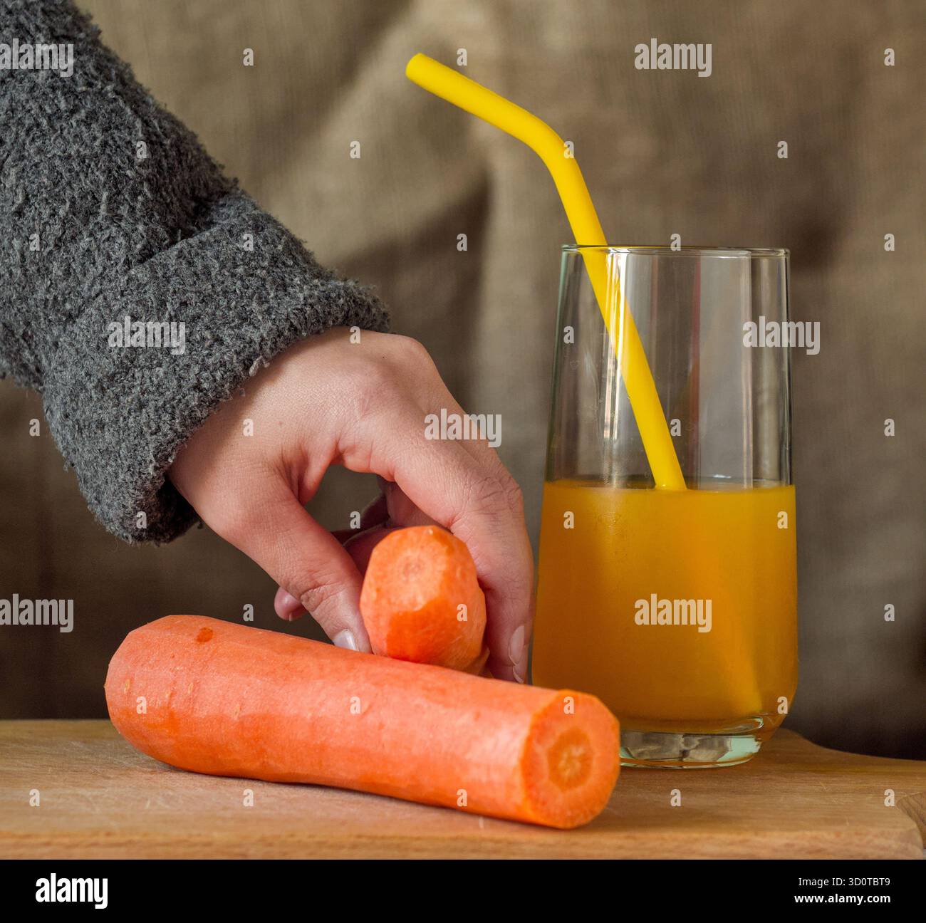 La mano di una persona prende una carota fresca su un tavolo accanto a un succo di arancia o di carota sano in bicchiere con paglia con sfondo burlap Foto Stock