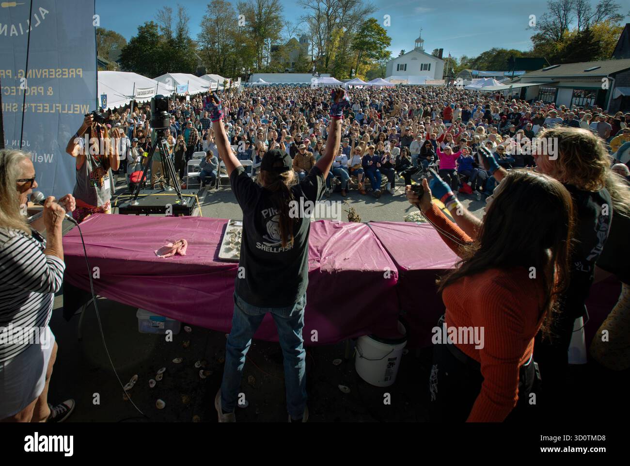 Wellfleet, Massachusetts, Stati Uniti. 18 ottobre 2025. Primo giorno del 25° OysterFest annuale di due giorni nella piccola città di Cape Cod, 100 miglia (160 km) via strada a sud-est di Boston, Massachusetts. Il festival sponsorizzato dalla Wellfleet Oyster Alliance (WOA), un'organizzazione no-profit che sostiene l'industria dei molluschi attrae oltre 20.000 visitatori nella piccola città di Cape Cod con una popolazione permanente di poco più di 3.500 abitanti. Foto: La folla fa il tifo a una donna che tiene in braccio il coltello dopo aver mosso le 24 ostriche grezze su un palco allestito nel parcheggio del municipio il primo giorno della gara. Foto Stock