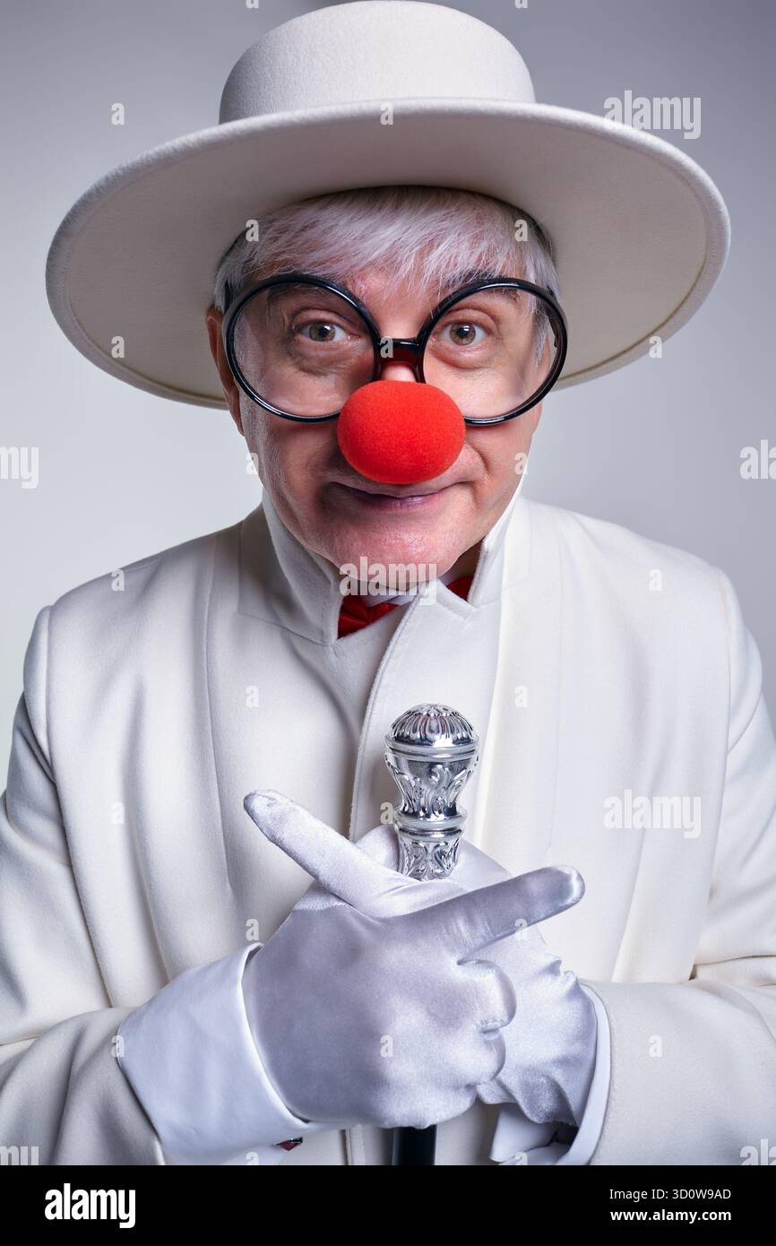 Un clown anziano con un cappotto bianco e un cappello con bastone da passeggio. L'uomo punta le dita in direzioni diverse. Foto Stock