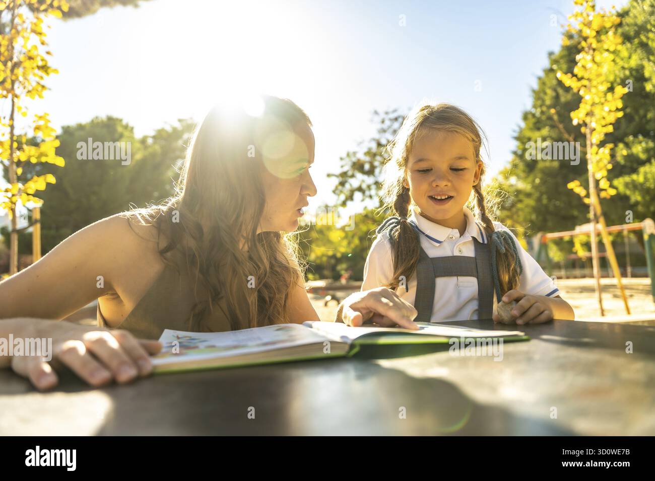 Madre e figlia leggono insieme un libro, seduti a un tavolo all'aperto in un ambiente scolastico luminoso e soleggiato, concentrandosi sull'istruzione e. Foto Stock