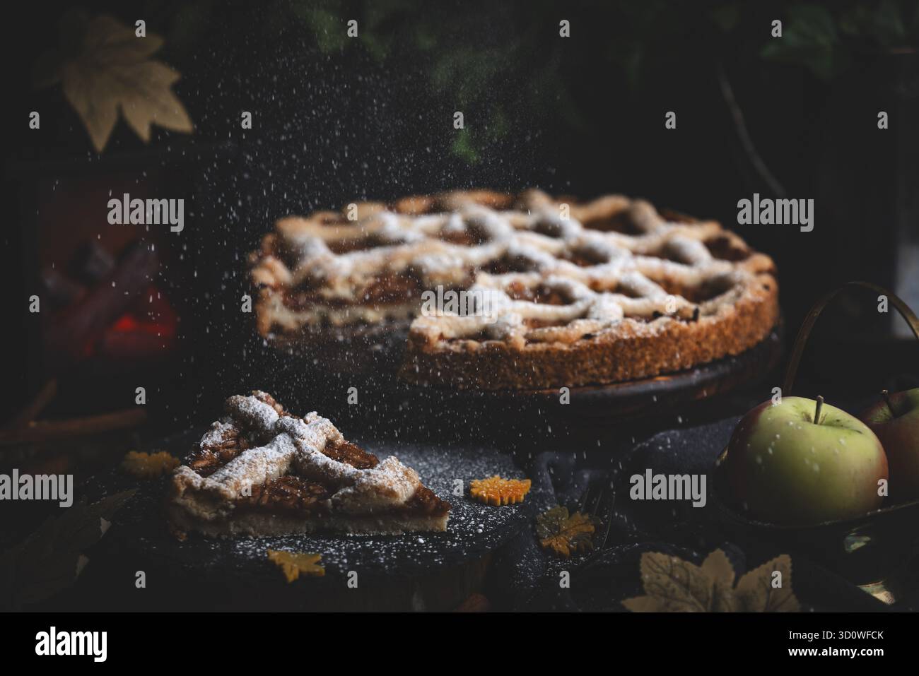 Lo zucchero a velo cade sulla torta di mele circondata da foglie autunnali in un ambiente buio Foto Stock