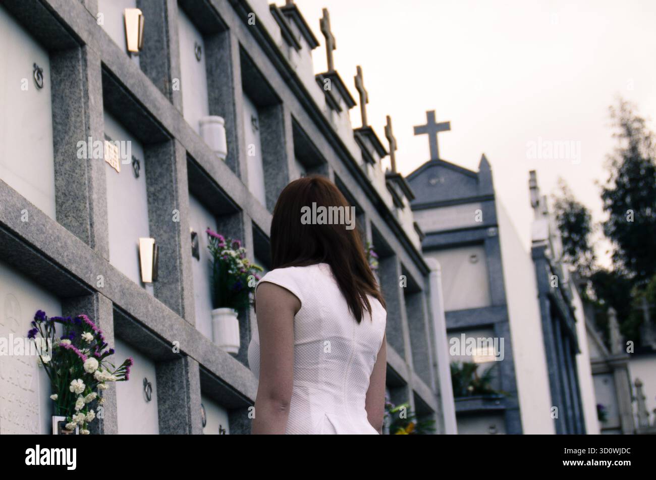Una donna vestita di bianco si erge tranquillamente davanti a un muro di tombe in un cimitero, circondato da croci e fiori. L'immagine trasmette perdita, memoria, Foto Stock