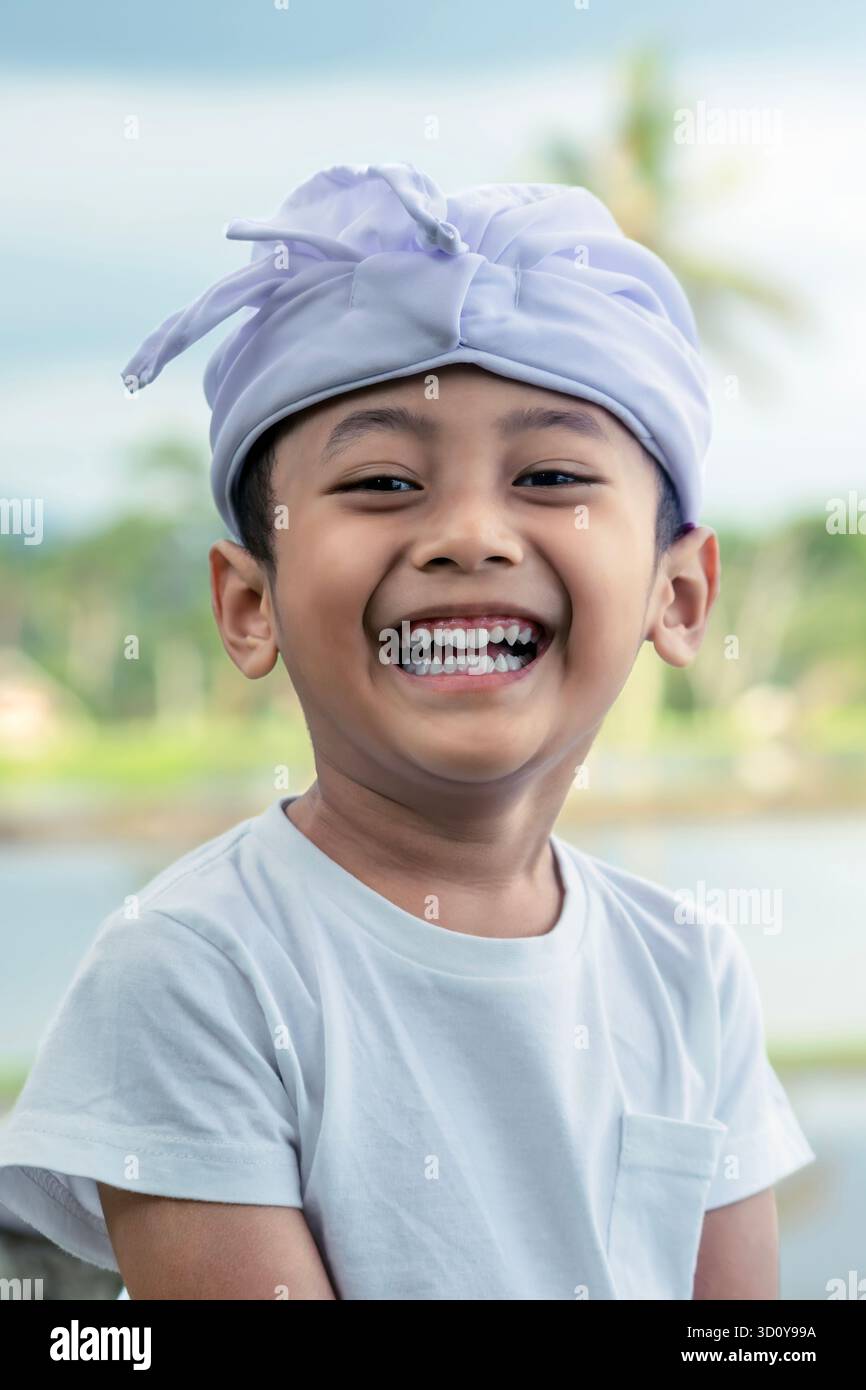 Ritratto ravvicinato di un bambino balinese che ride gioiosamente con la fascia cerimoniale bianca "Udeng" con un campo di riso sfocato sullo sfondo il giorno delle Galungan. Foto Stock