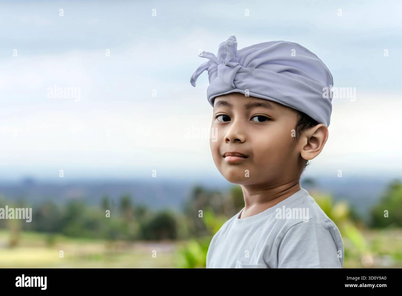 Ritratto di un bambino balinese con fascia e camicia cerimoniali bianche "Udeng", in piedi all'aperto con un campo di riso sfocato sullo sfondo il giorno delle Galungan. Foto Stock