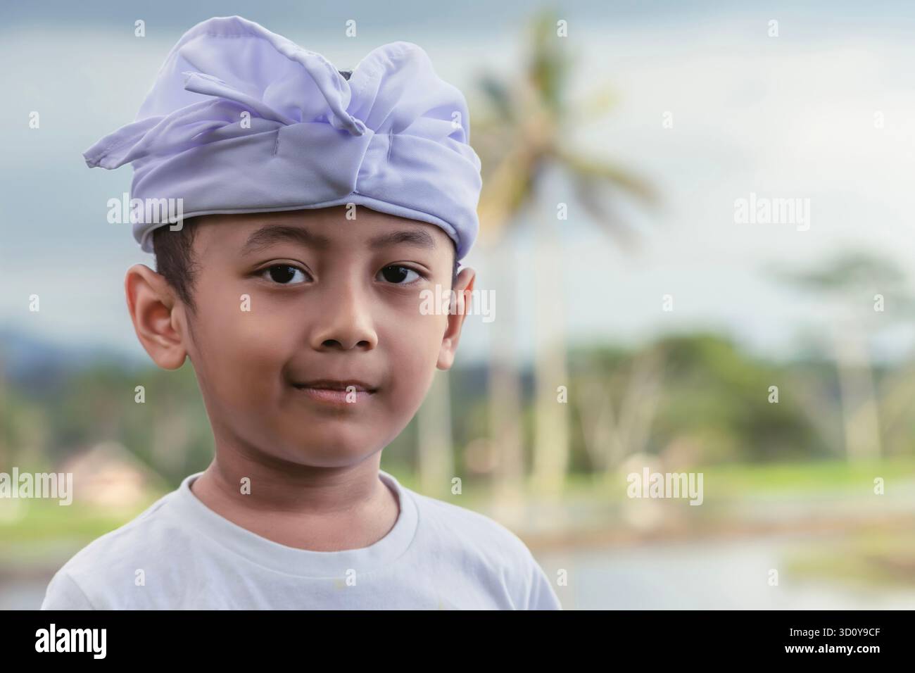Ritratto di un bambino balinese con un sorriso fresco e sottile in abiti cerimoniali bianchi e un campo di riso leggermente sfocato sullo sfondo il giorno delle Galungan. Foto Stock