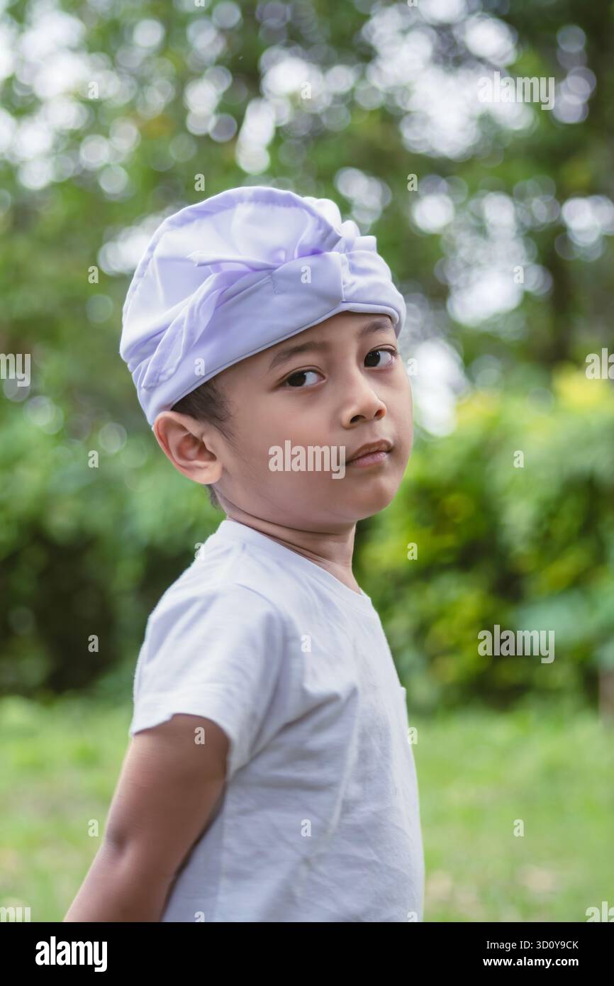 Ritratto verticale di un bambino balinese che indossa un cerchietto cerimoniale bianco "Udeng", incorniciato contro la vegetazione sfocata del giardino. Foto Stock