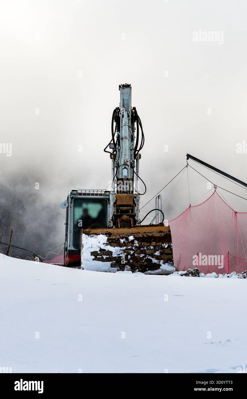 Un cantiere innevato con un escavatore. La macchina è posizionata su un pendio, circondata da un'atmosfera nebbiosa e da una rete di sicurezza rossa. Foto Stock