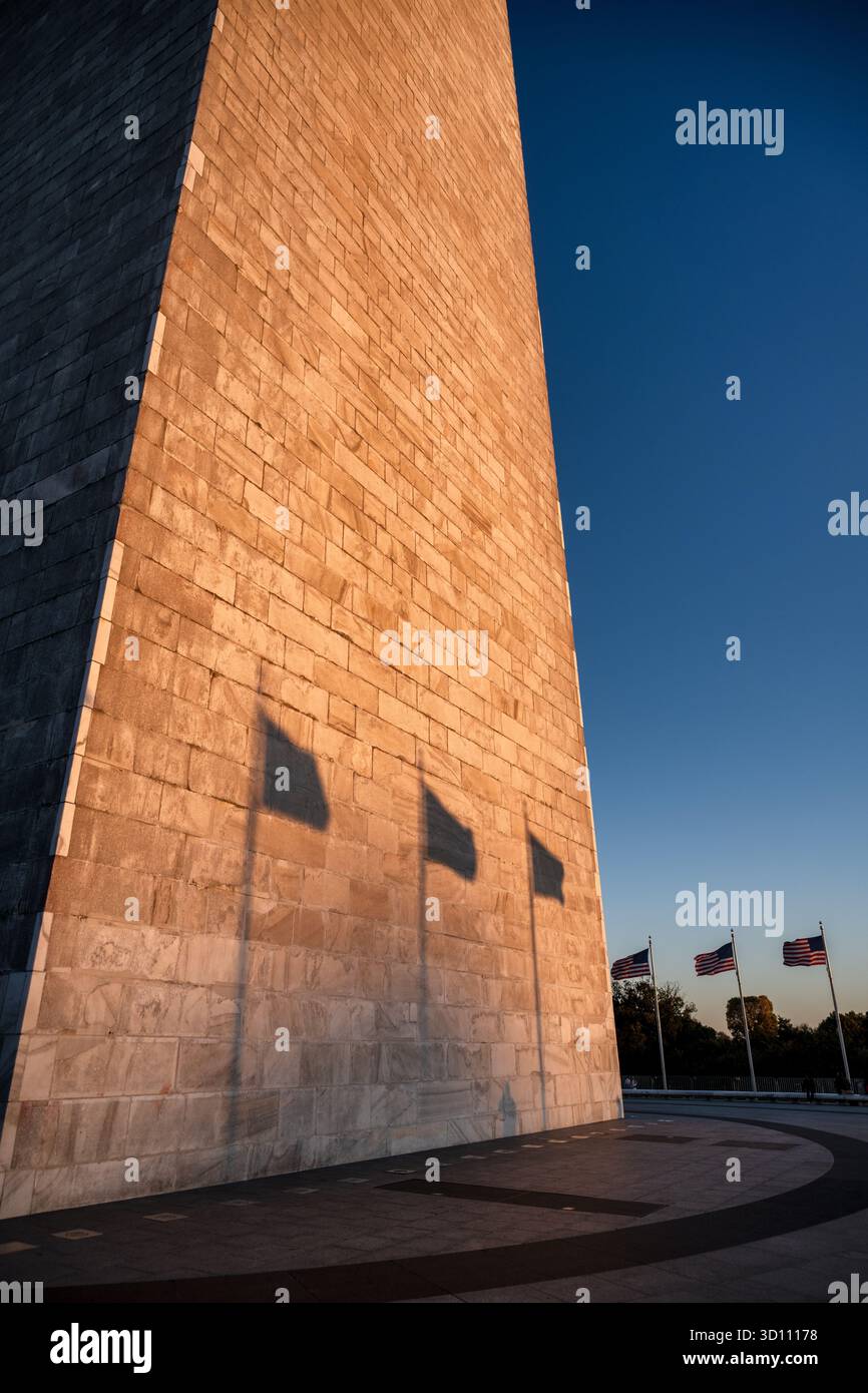 Monumento a Washington al tramonto con la bandiera delle Ombre Washington DC // WASHINGTON DC - il monumento a Washington è visibile al tramonto, con il sole basso che getta lunghe ombre di bandiere americane sulla sua facciata in pietra. Questo iconico obelisco, costruito in onore di George Washington, è un importante punto di riferimento sul National Mall. Costruito in marmo, granito e gneiss di pietra blu, raggiunge un'altezza di 554 piedi 7 pollici (169,046 metri). Cinquanta bandiere americane circondano la base del monumento, rappresentando gli stati. Il sole che tramonta bagna un lato della struttura in una calda luce dorata. Foto Stock