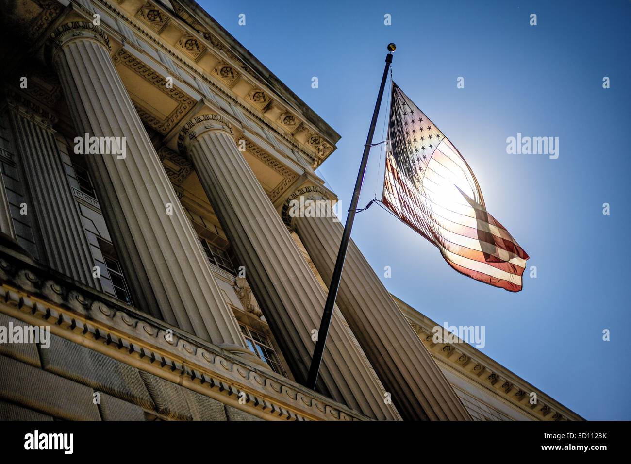 Bandiera DEGLI STATI UNITI retroilluminata dal sole sull'Herbert C Hoover Federal Building Washington DC // WASHINGTON DC - la bandiera degli Stati Uniti è retroilluminata dal sole sull'Herbert C. Hoover Federal Building, sede del Dipartimento del commercio degli Stati Uniti. Situata in Pennsylvania Avenue NW a Washington DC, questa struttura monumentale è una componente chiave del Triangolo Federale. Progettato in stile neoclassico dall'architetto Waddy B. Wood, l'edificio è stato costruito tra il 1928 e il 1932. La sua grande facciata e la sua posizione prominente riflettono il suo ruolo nella capitale della nazione. Foto Stock