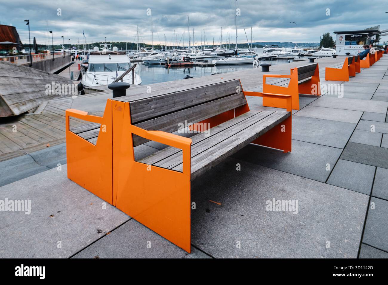 Oslo, Norvegia - 8 settembre 2025: Panche vuote in legno color arancio su argini e barche ormeggiate nel porto del distretto di Aker Brygge Foto Stock