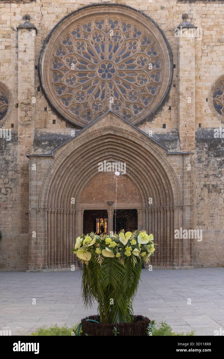 La facciata della chiesa in pietra presenta una grande rosetta, ingresso ad arco e una fontana decorativa con fiori e palme Foto Stock