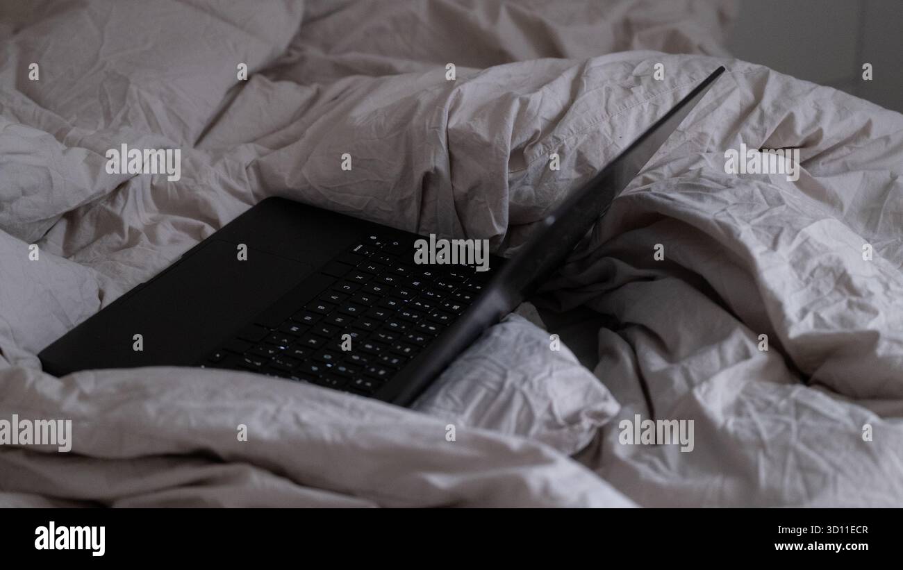 Laptop nero su biancheria da letto non fatta di luce, fotografato con luce naturale soffusa. L'ambiente accogliente riflette il lavoro remoto, il relax e lo stile di vita domestico. Ideale f Foto Stock