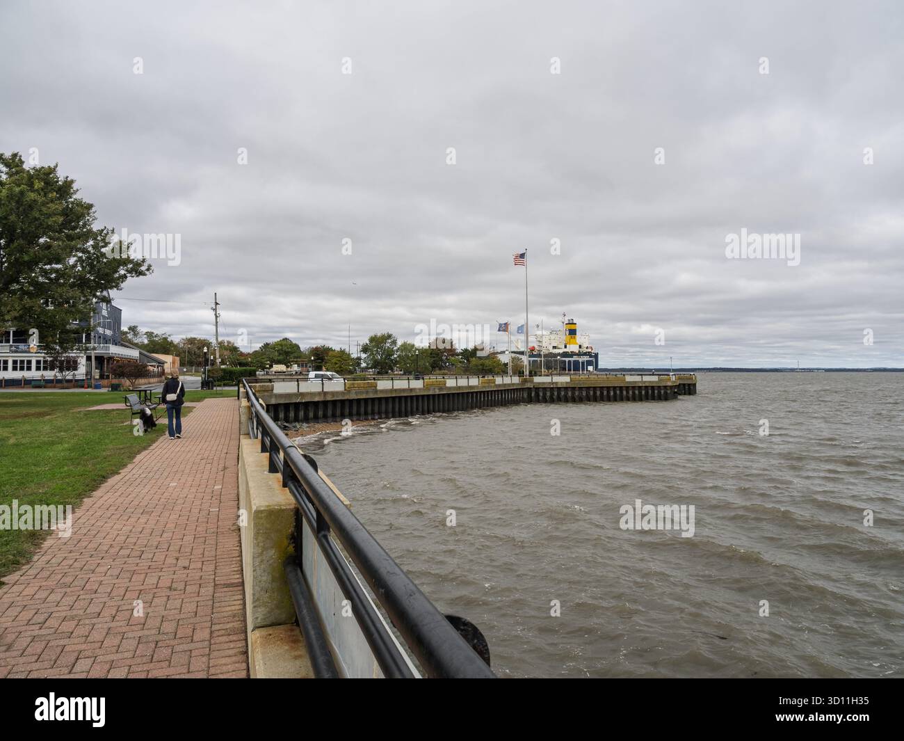 Passeggiata in mattoni e molo a Delaware City con una lontana nave da carico sulle ampie e grigie acque del fiume Delaware. Foto Stock