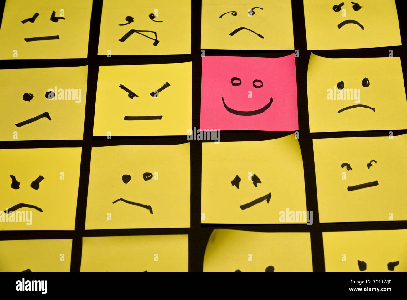 15 carte gialle poste l'una parallelamente all'altra con facce disegnate su di esse che esprimono rabbia o tristezza, di fronte ad una carta rosa con un volto che esprime felicità Foto Stock