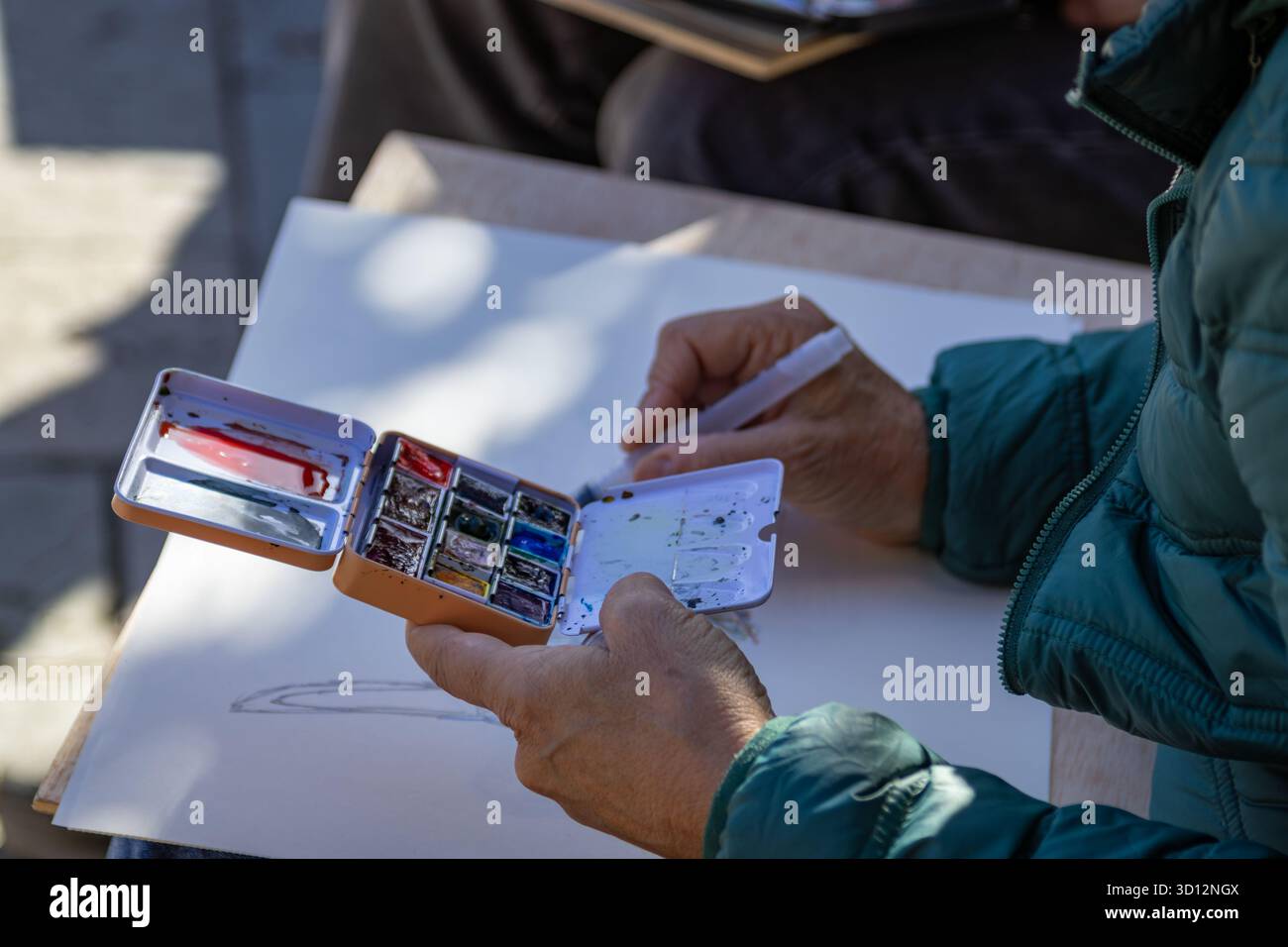 Artista che utilizza un pennello ad acqua e una tavolozza compatta di acquerelli su carta bianca per esterni. Concetto di hobby creativo Foto Stock