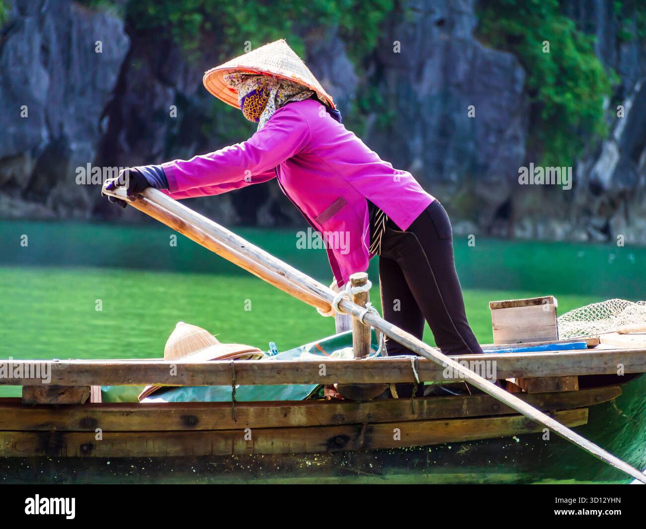 Canottaggio tradizionale in vivace abito viola ao dai e cappello conico che rema in sampan di legno attraverso le acque color smeraldo della baia di ha Long, Vietnam Foto Stock