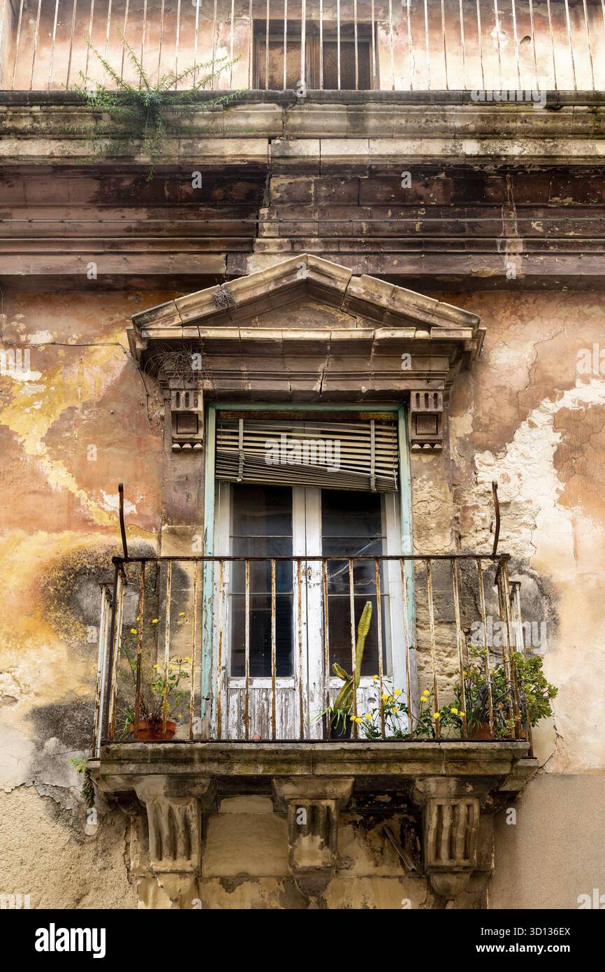 Ragusa, Sizilien - Italia - 04-03-2025: Balcone in pietra con ringhiere arrugginite, piante in vaso e facciata sbucciante su un vecchio edificio a Ragusa, Sici Foto Stock