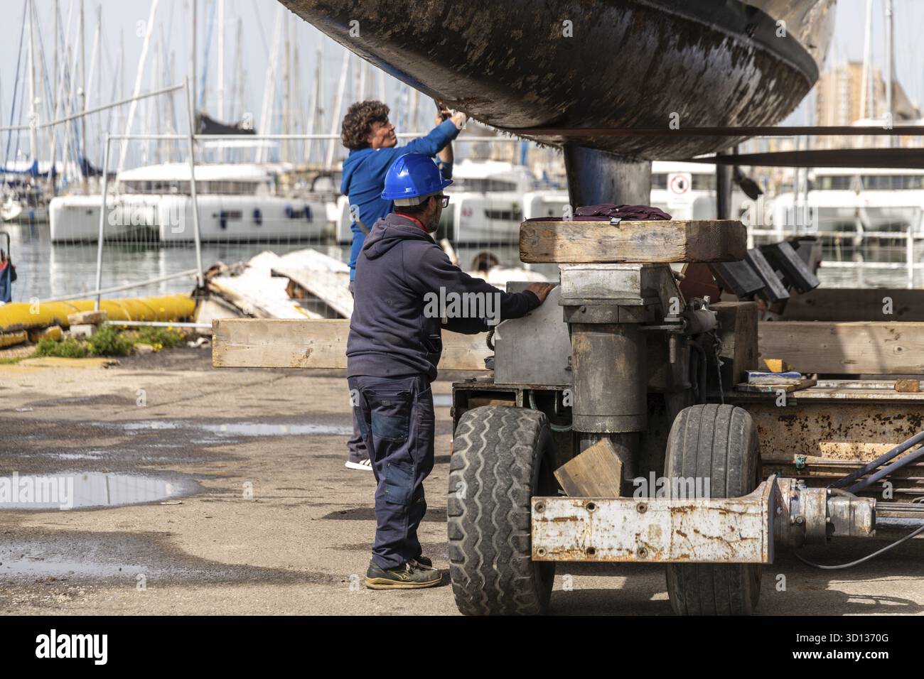 Cagliari - Sardinien - Italien - 03-14-2025: Due operai esaminano ed eseguono la manutenzione sul lato inferiore di un catamarano sollevato su un rimorchio vicino a un ma Foto Stock