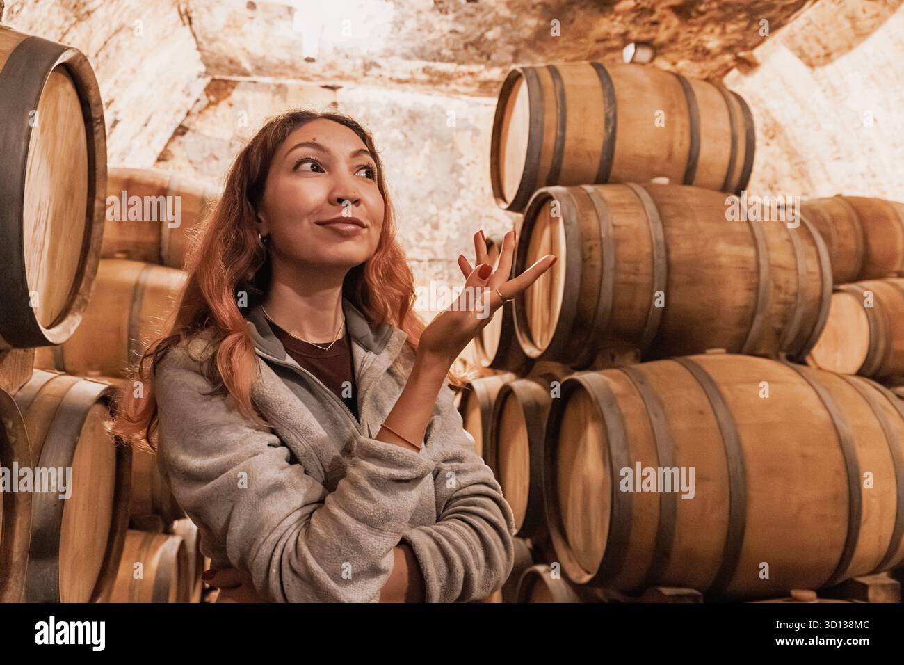 Woman si erge tra botti di rovere impilate in una cantina tradizionale, esplorando la produzione di vino e degustando il turismo Foto Stock
