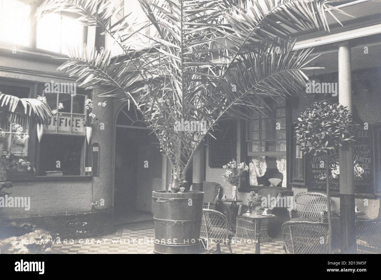 Una torreggiante palma fissa il cortile del Great White Horse Hotel, Ipswich, circondato da sedie in vimini e pavimento piastrellato. Le finestre con tende in pizzo e la segnaletica incorniciano lo spazio, mentre la finestra "Office" suggerisce la reception nelle vicinanze. Pubblicata intorno al 1910, questa cartolina cattura l'ospitalità edoardiana con un tocco tropicale in una delle strutture più storiche del Suffolk. Da una cartolina originale non attribuita. Foto Stock