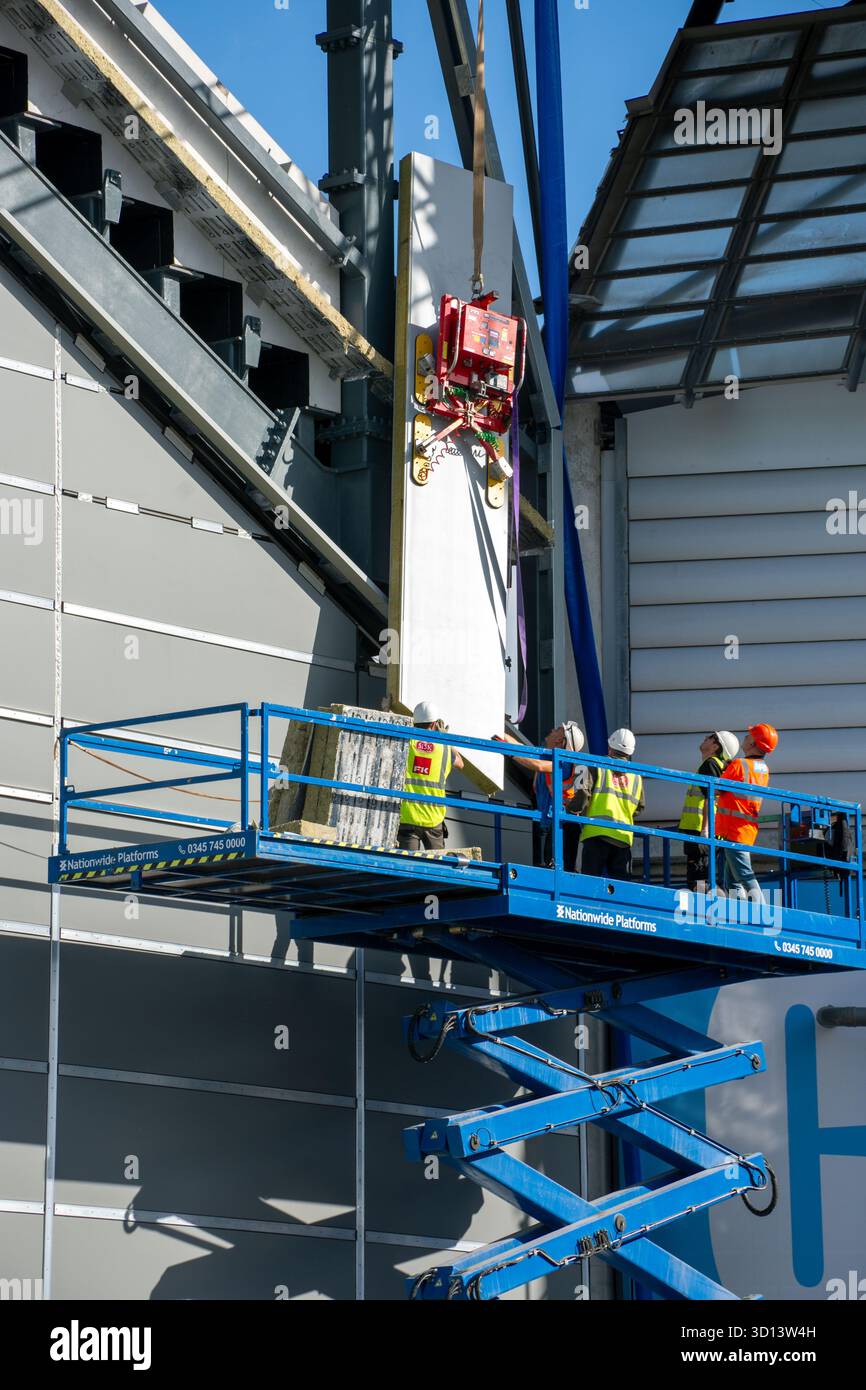 Lavoratori su una piattaforma di accesso che montano una lastra di rivestimento utilizzando un sollevatore a ventosa su una gru. Etihad North Stand Expansion, Manchester, Inghilterra, Regno Unito. Foto Stock