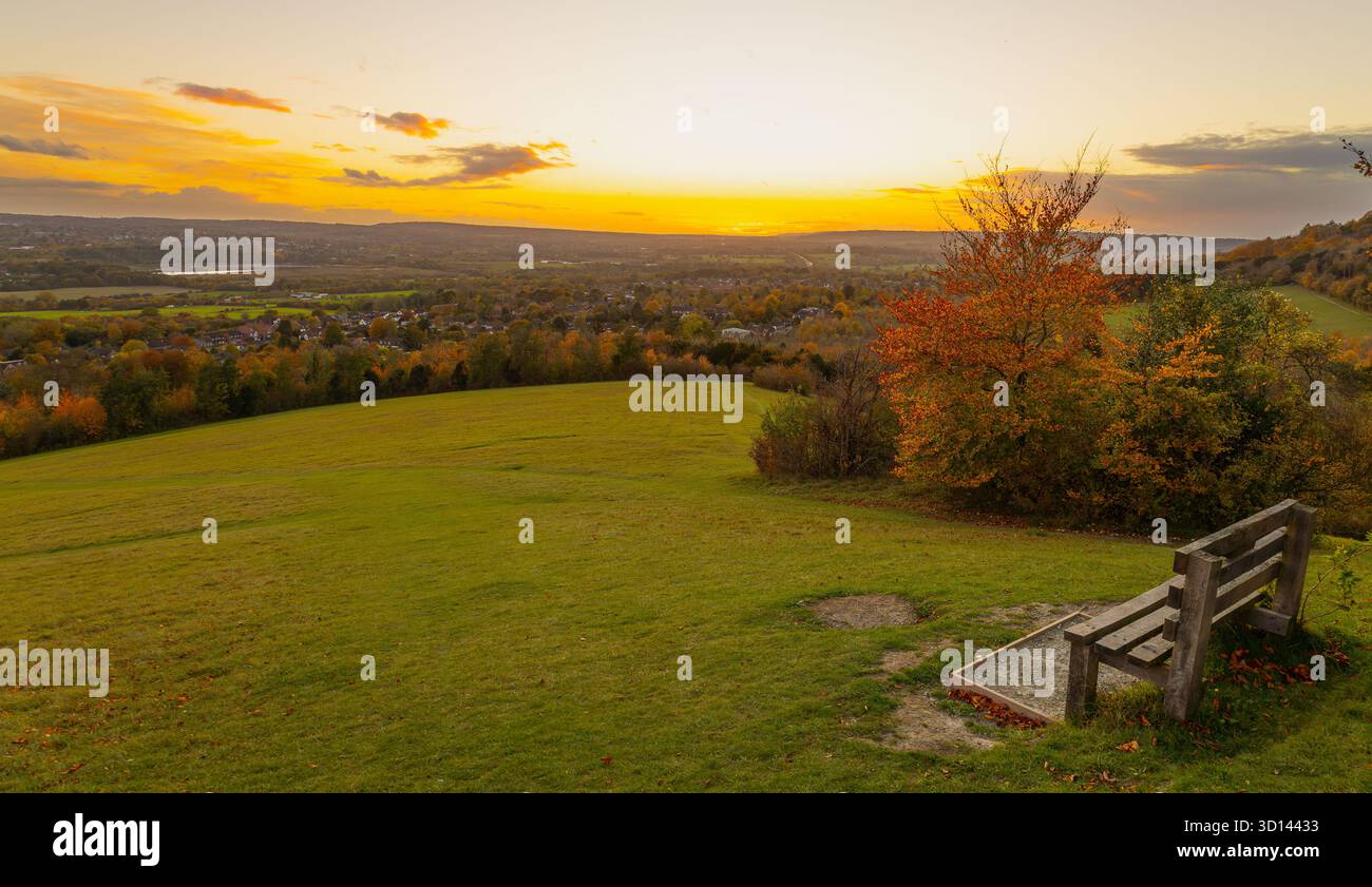 Banco singolo vuoto in cima a Kemsing Downs che guarda verso il tramonto su Kemsing e Otford nel Kent. Alberi e colline con colori autunnali e cielo vibrante Foto Stock