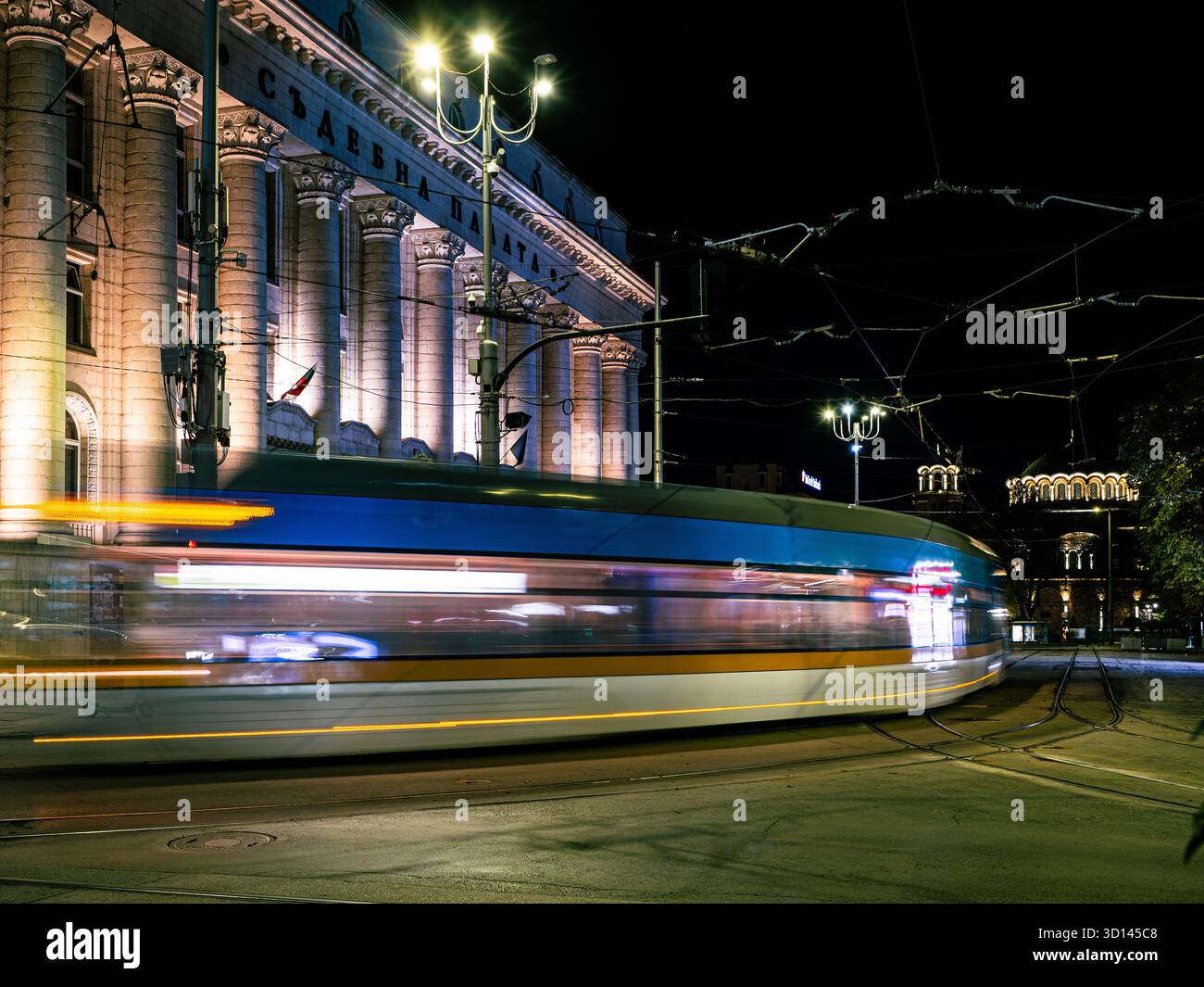 Sofia, Bulgaria - 24 ottobre 2025: Una lunga esposizione cattura un tram cittadino che passa accanto alle colonne illuminate dello storico tribunale di Sofia (Sadebna) Foto Stock