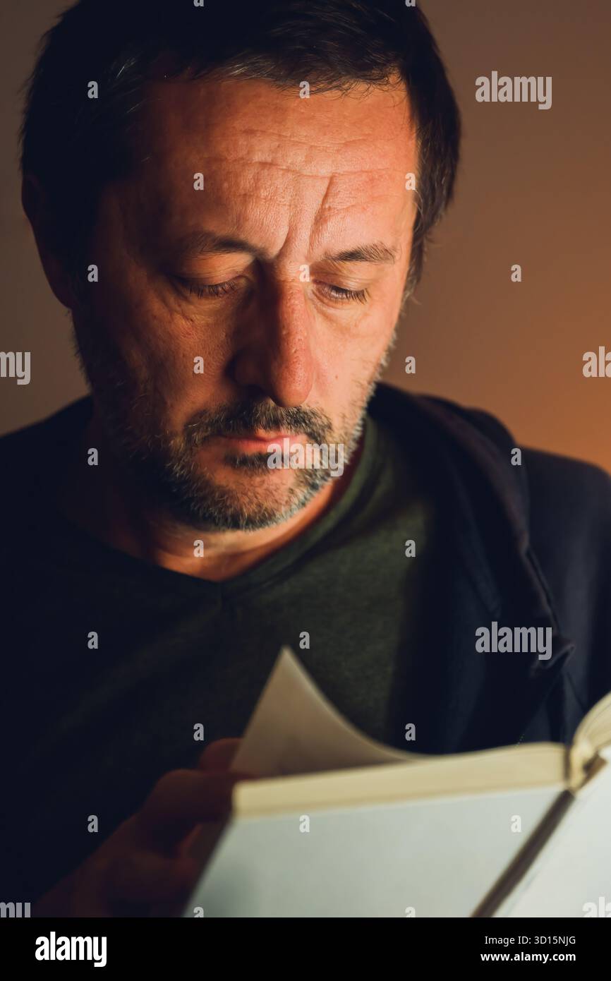 Ritratto basso di un uomo maturo immerso in un libro sotto una luce calda, catturando solitudine, messa a fuoco e una contemplazione intellettuale silenziosa. Messa a fuoco selettiva. Foto Stock