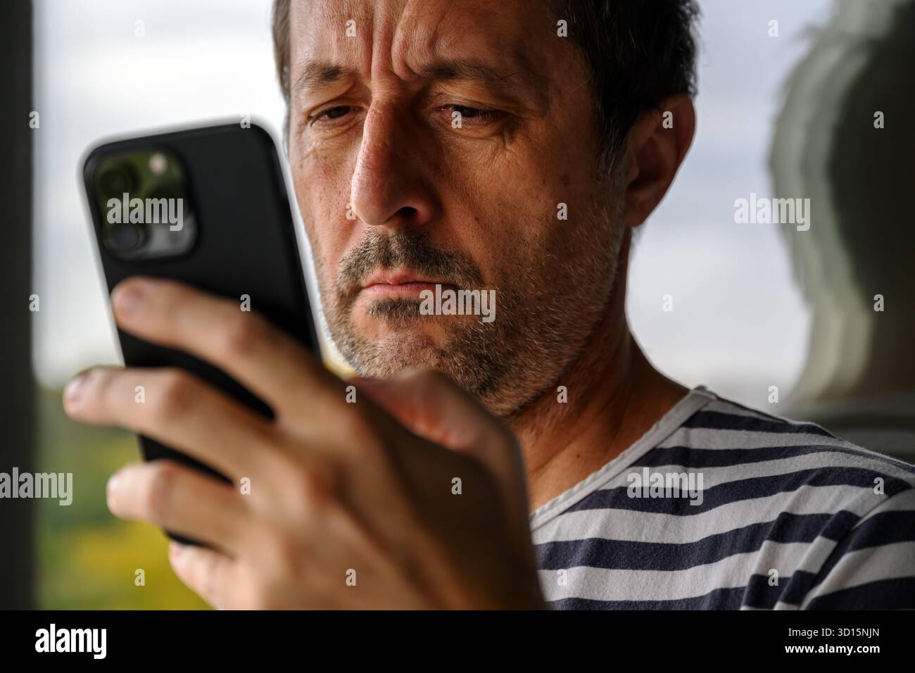 Uomo maturo in camicia a righe da marinaio con smartphone sul balcone dell'appartamento, riflesso in vetro, concetto di comunicazione e stile di vita. F. Selettiva Foto Stock