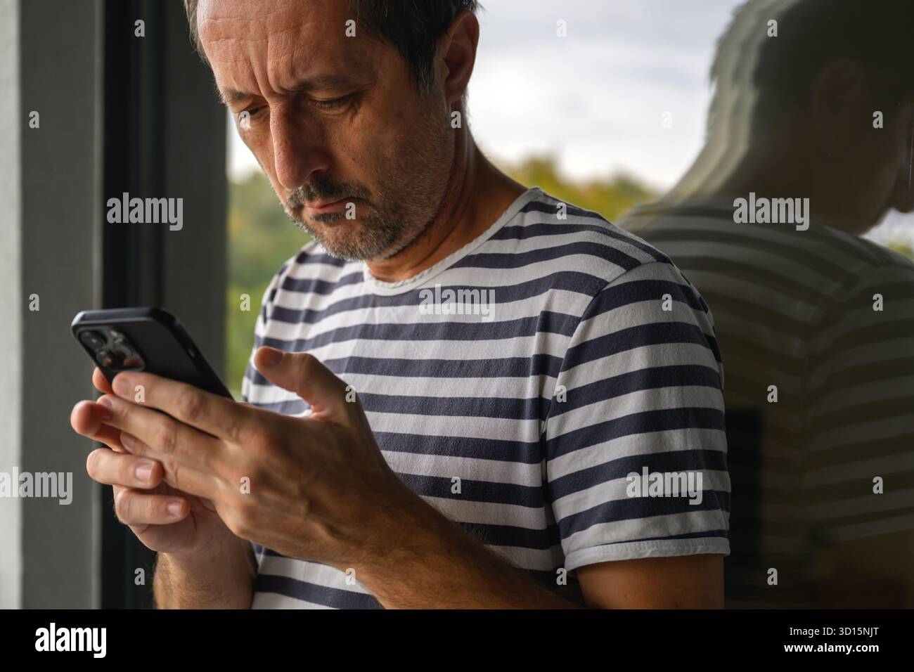 Uomo maturo in camicia a righe da marinaio in piedi sul balcone e scrivendo su smartphone, riflesso in vetro della finestra, simboleggia la comunicazione moderna e soli Foto Stock