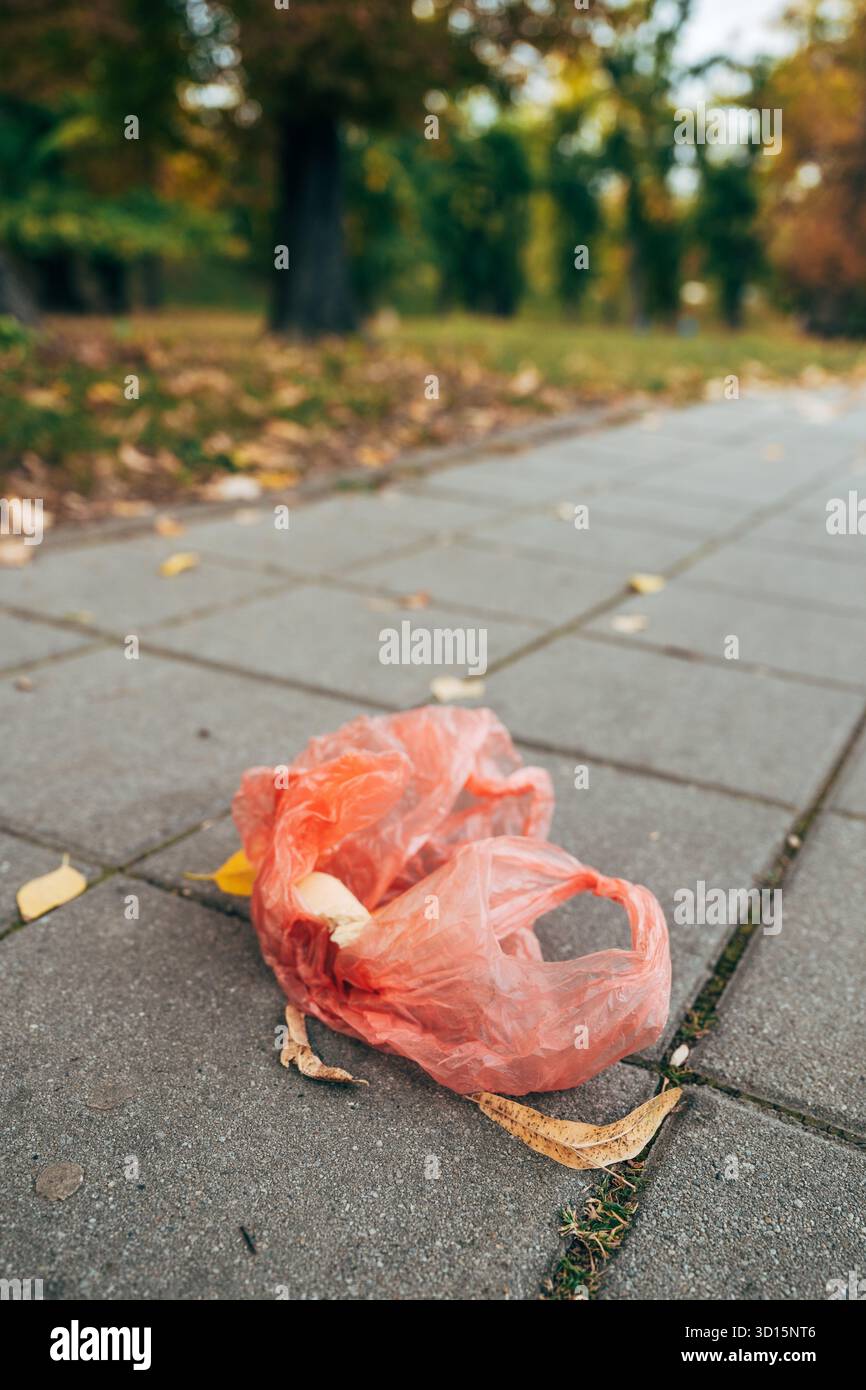 Sacchetto della spesa di plastica rossa gettato adagiato sul marciapiede del parco, che simboleggia i rifiuti urbani e l'inquinamento ambientale, evidenziando i rifiuti e le persone Foto Stock