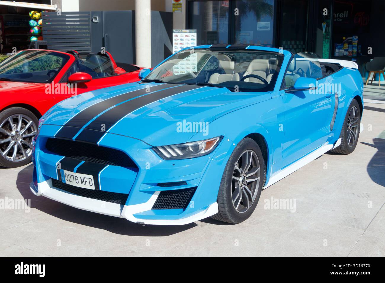 Auto sportiva Ford Mustang convertibile blu brillante con strisce nere da corsa parcheggiata di fronte a un moderno showroom in una giornata di sole Foto Stock