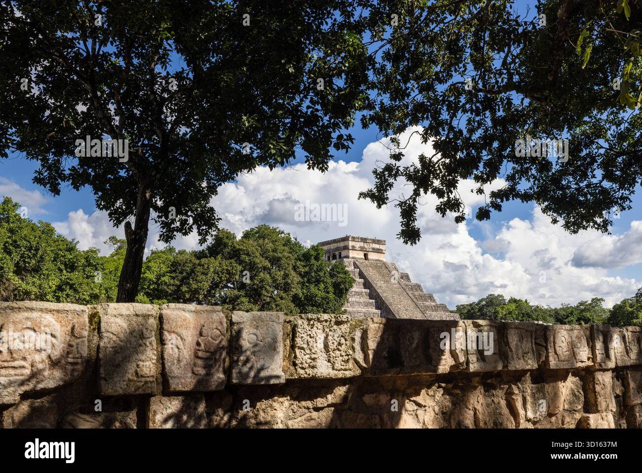 Piramide di El Castillo incorniciata da alberi con intagli al teschio di Tzompantli in primo piano, Chichén Itzá. Foto Stock