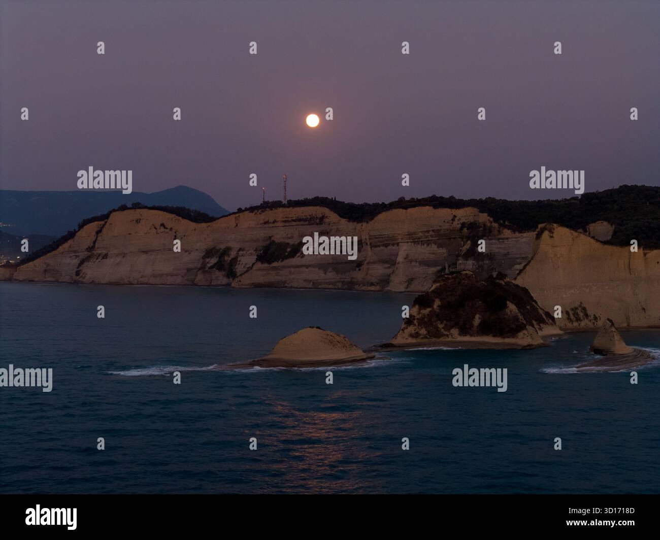 Una luminosa luna piena sorge in un cielo viola crepuscolo sopra le spettacolari scogliere bianche e le montagne marine di Capo Drastis, Corfù, con il suo riflesso sul cal Foto Stock