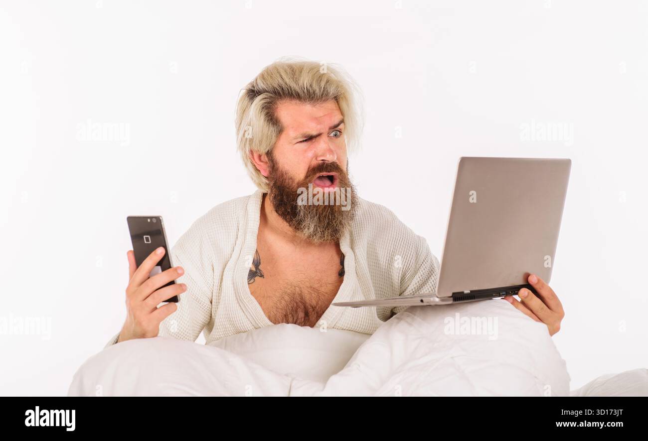 Lavora da casa. Uomo che lavora al computer portatile a letto la mattina. Uomo d'affari furioso con smartphone e laptop. Uomo barbuto seduto sul letto e che lavora al portatile. Foto Stock