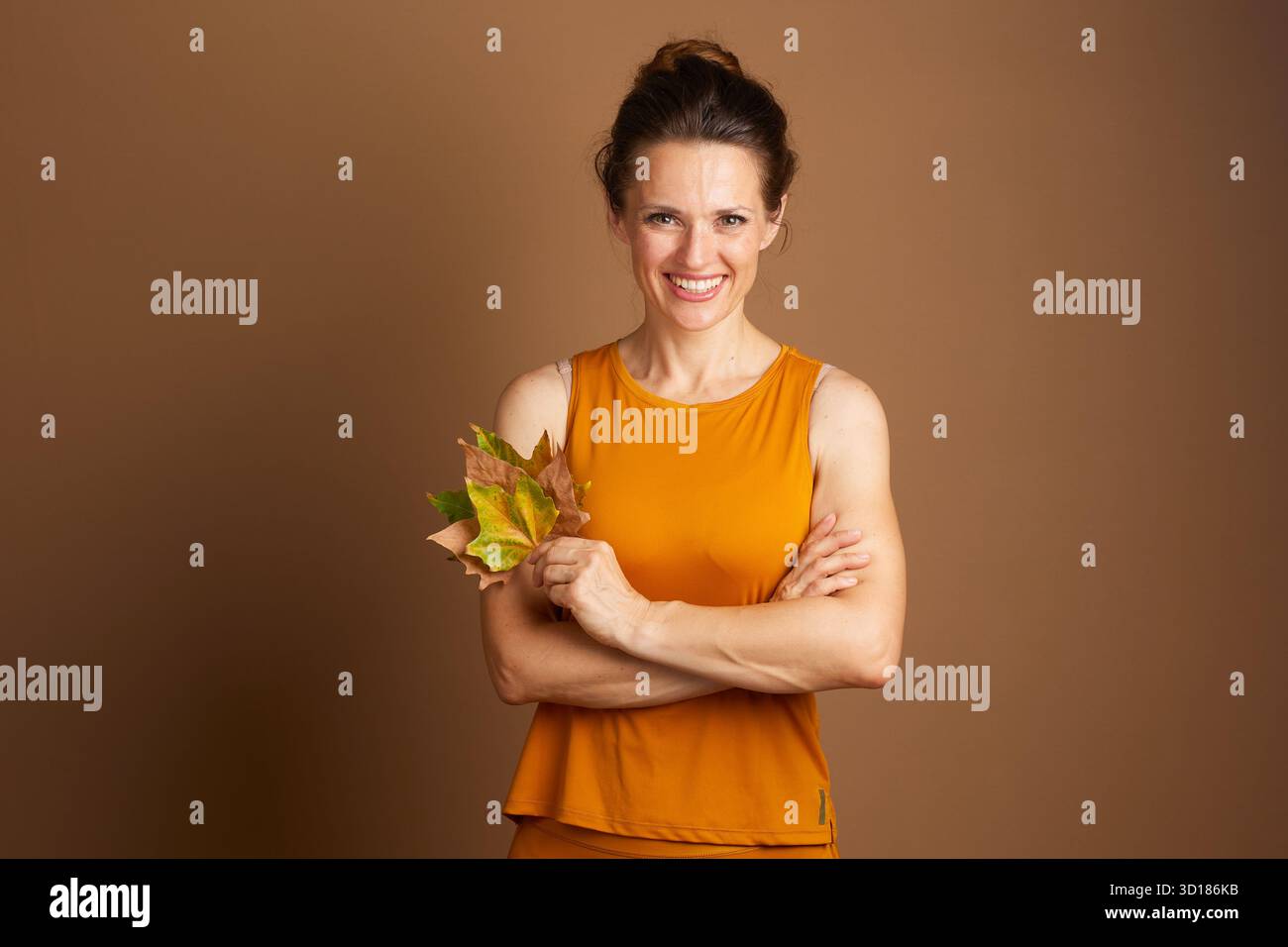 Una donna adulta sorridente con capelli scuri, con canotta color senape, si stende con le braccia incrociate, tenendo le foglie autunnali su un dorso marrone pieno Foto Stock