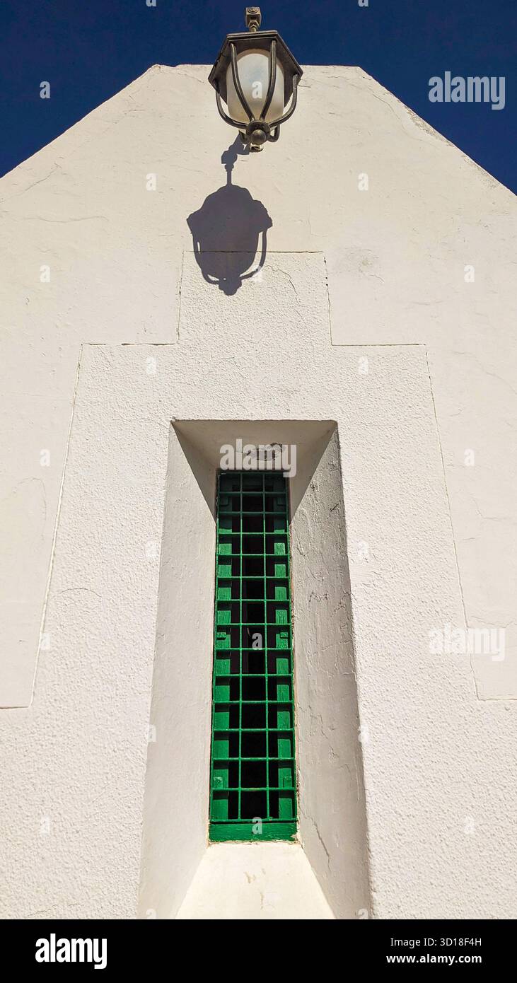 Dettagli architettonici minimalisti di una parete dipinta di bianco con una stretta finestra barrata verde e una lanterna vintage che getta una forte ombra sotto il cielo blu - Immagine stock catturata con smartphone